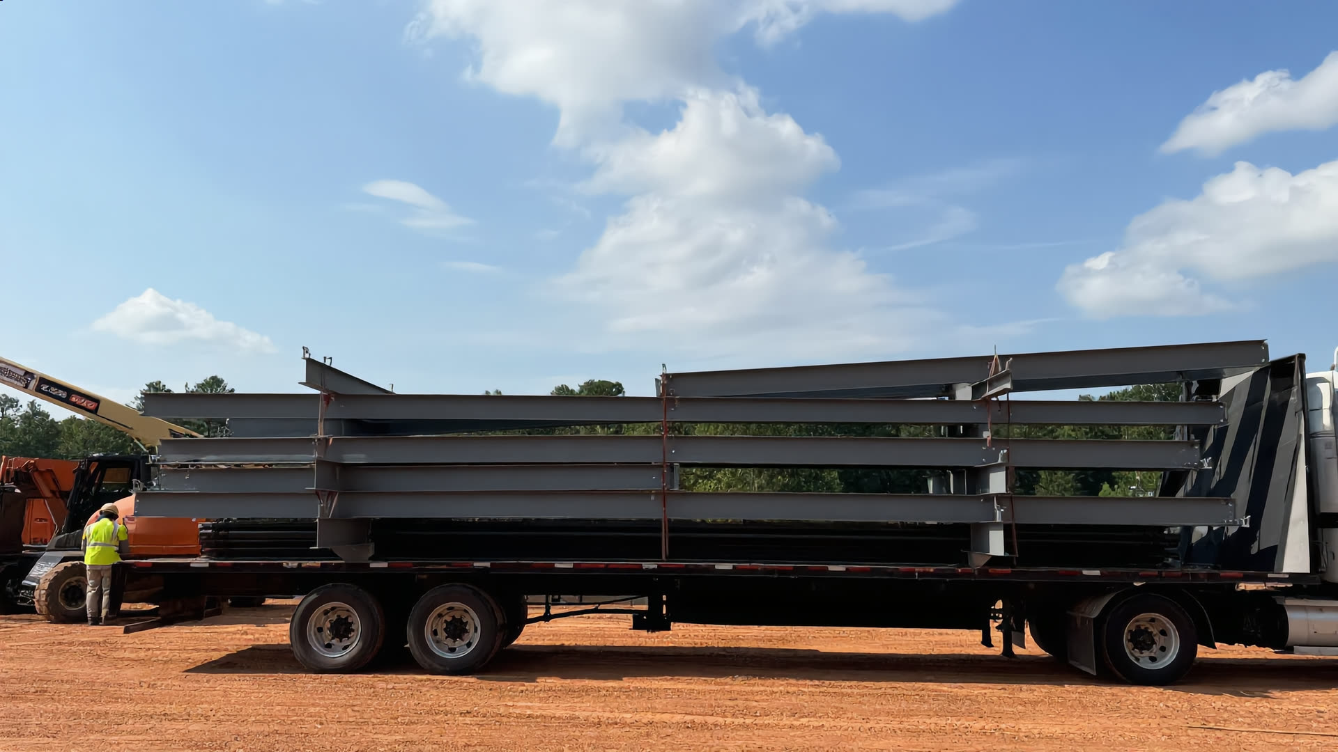 A flatbed trailer loaded with fabricated steel beams standing on red Georgia clay at a jobsite