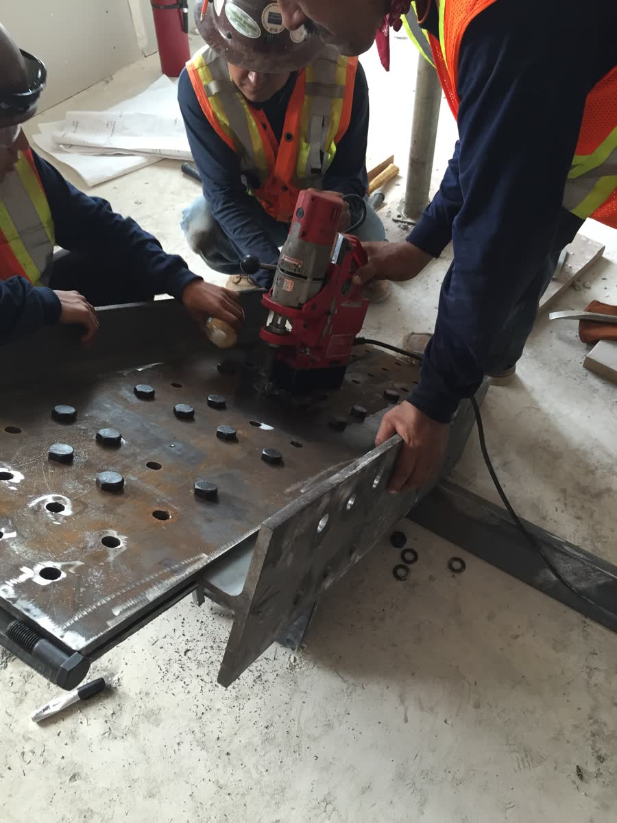 Three More Steel crew members in hard hats and high-visibility vests drilling a heavy steel base plate